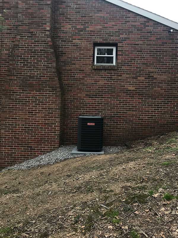 Heat pump condenser on side of home. The yard is sloped and the chimney is to the left.