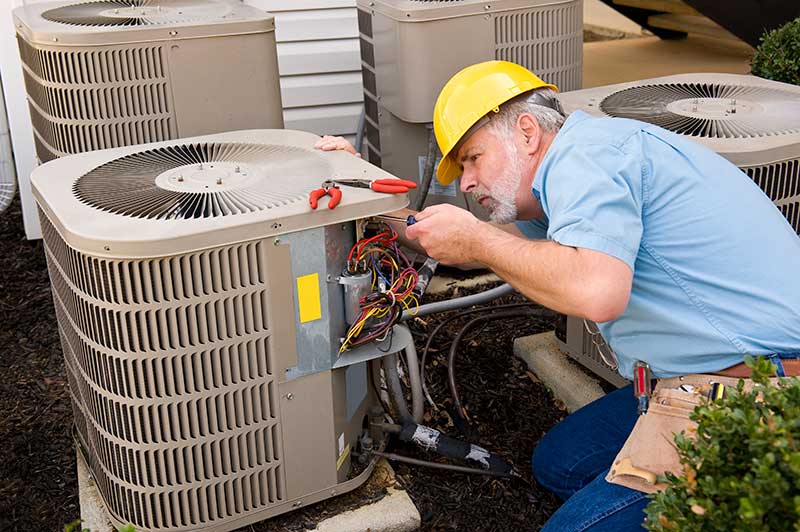 Technician working on Air Conditioner wearing a hard hat. He has a gray beard and mustache. He has guages hooked up to different color wires inside the unit.
